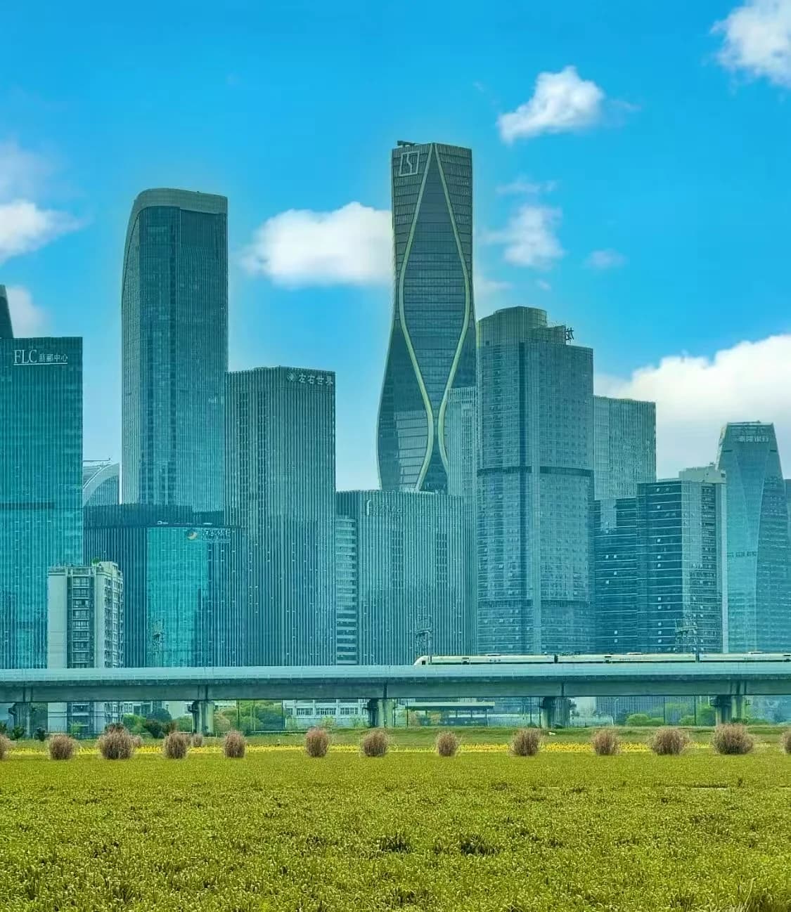 Modern towers rise behind a green field and an elevated railway
