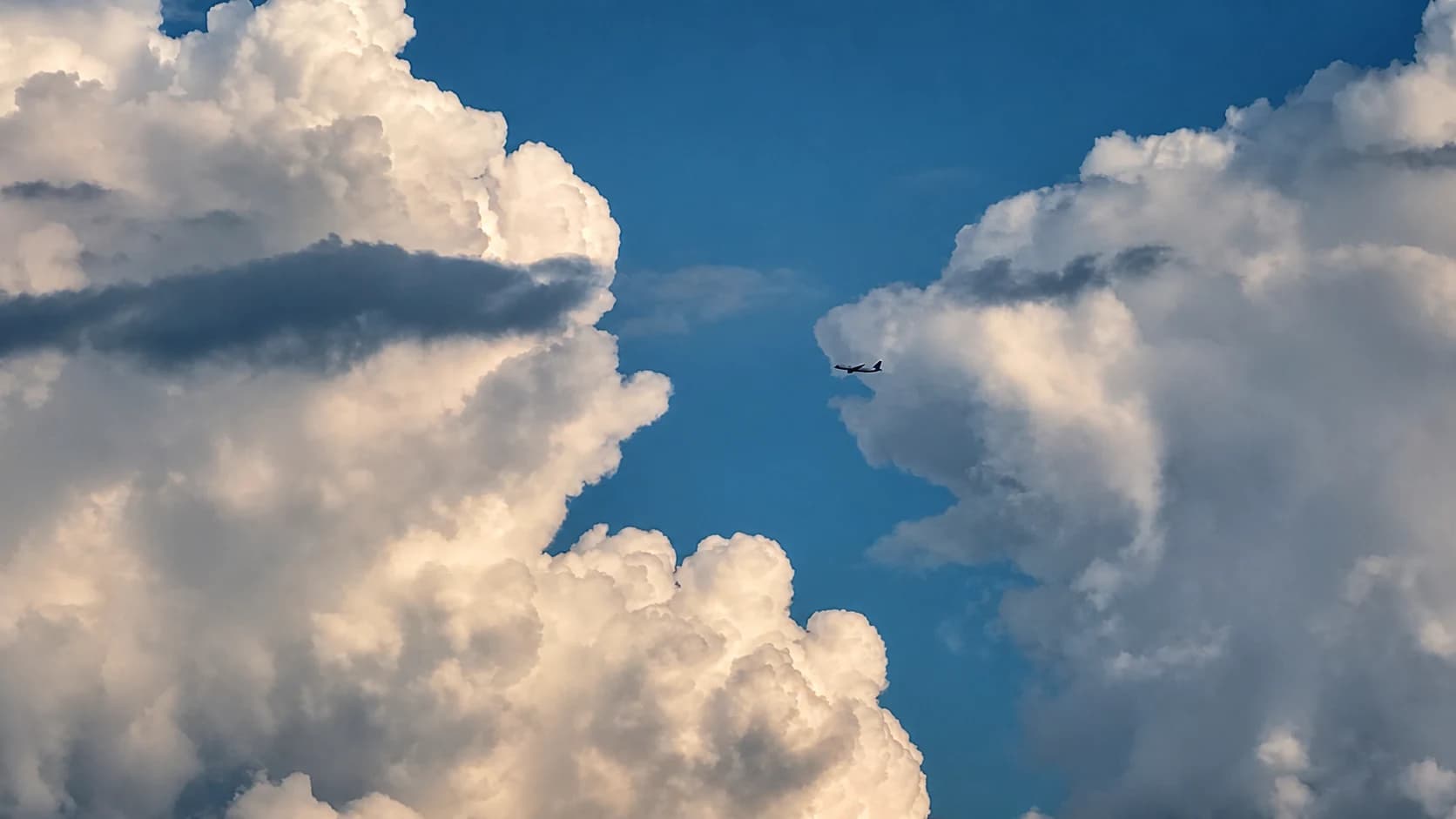 A small airplane passes between towering white clouds in a deep blue sky